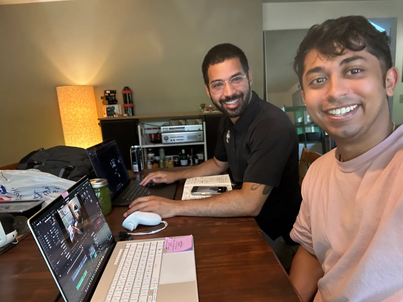 Selfie of Sid grinning in the foreground while Morgan laughs at a desk holding a Quest controller, laptops and sticky notes around them